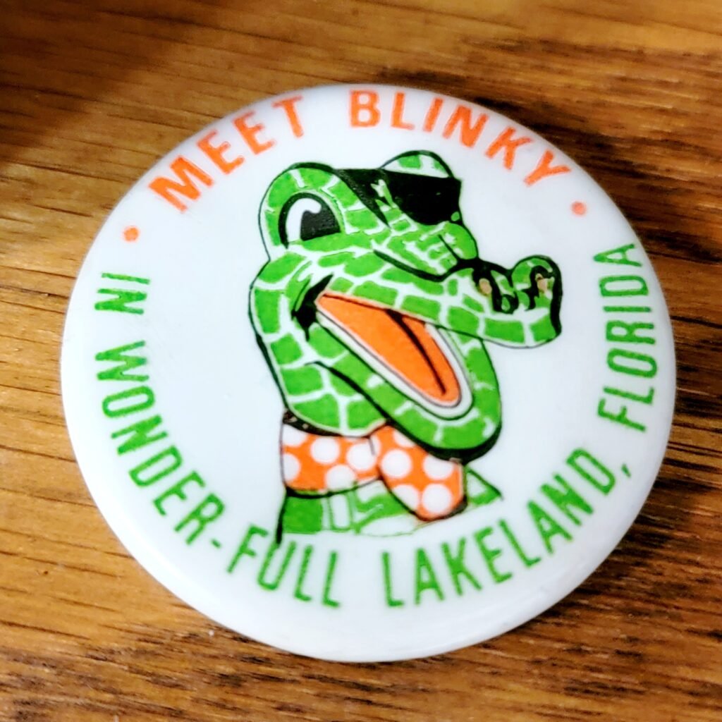 Vintage 'Meet Blinky' pinback button featuring Blinky the one-eyed alligator, a famous attraction from Lakeland, Florida. The design showcases a cartoon-style green alligator wearing an eye patch, sunglasses, and orange polka-dot swim trunks, surrounded by the text 'Meet Blinky' in orange and 'In Wonder-Full Lakeland, Florida' in green. A nostalgic piece of Florida roadside attraction history.