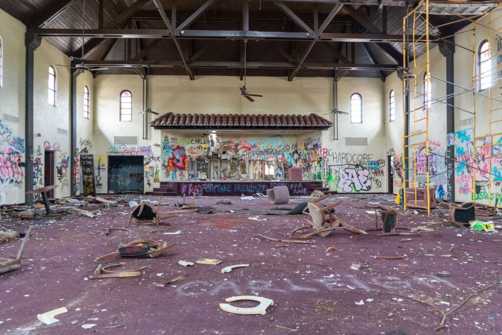 Interior of the abandoned Port of the Islands Hotel in Naples, FL, now demolished, showcasing a large, graffiti-covered room in disrepair. Debris is scattered across the floor alongside broken furniture, and the walls feature prominent graffiti. Exposed wooden beams line the ceiling, and light streams through several arched windows, creating a stark contrast with the state of decay. A scaffold is visible on the right, hinting at possible renovation or demolition work. This evocative scene captures the haunting atmosphere and deterioration of the historic site."