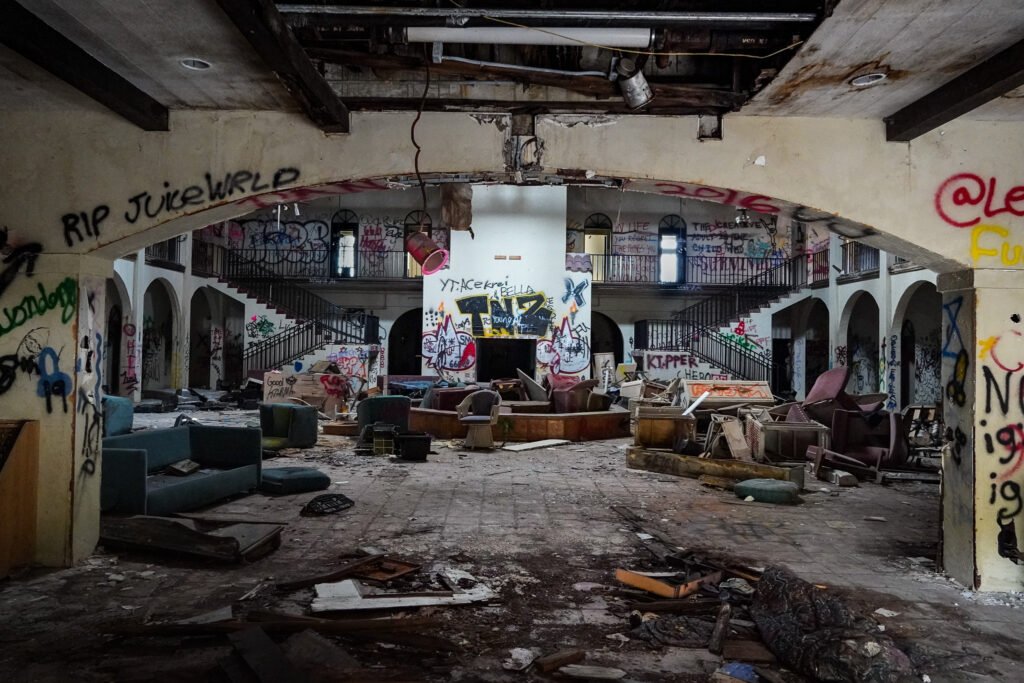 Abandoned Port of the Islands Resort in Naples, Florida, showing a decaying hotel lobby covered in graffiti. The once-luxurious space is now in ruins, with broken furniture, scattered debris, and crumbling ceilings. The grand staircase and arched walkways hint at the hotel's former elegance, now overtaken by urban exploration and vandalism. Graffiti tags, including 'RIP Juice WRLD' and various other markings, cover the walls. This eerie and abandoned location has become a hotspot for urban explorers and photographers documenting Florida’s forgotten places.