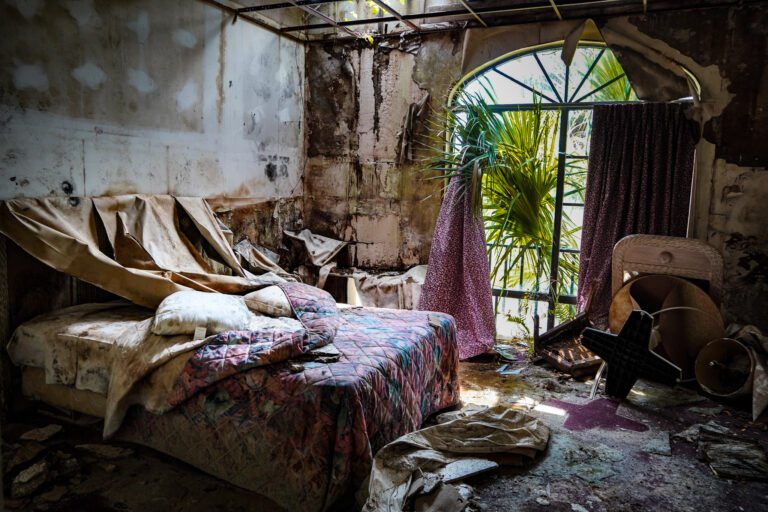 Decaying, abandoned room with a bed and broken furniture, showcasing neglect and urban decay. The image features a tattered bed with dirty bedding, peeling walls, a broken ceiling, and a large window with shattered glass and torn curtains revealing overgrown plants outside. Debris and damaged items, including a broken fan, litter the floor. Perfect for illustrating themes of urban exploration, abandonment, or the passage of time.