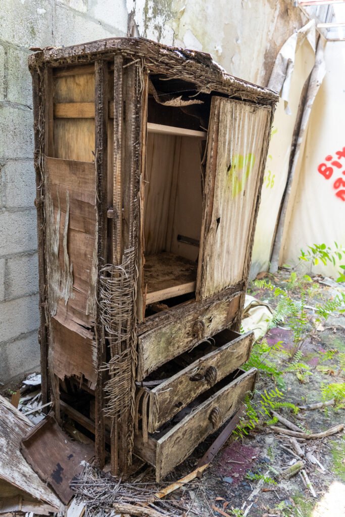 A heavily decayed wooden wardrobe inside the abandoned Port of the Islands Resort in Naples, Florida. The structure is falling apart, with broken drawers, frayed wood, and peeling veneer, surrounded by debris and overgrown vegetation. This eerie scene highlights the effects of time and nature reclaiming the once-luxurious Everglades hotel, making it a fascinating location for urban exploration and abandoned photography.