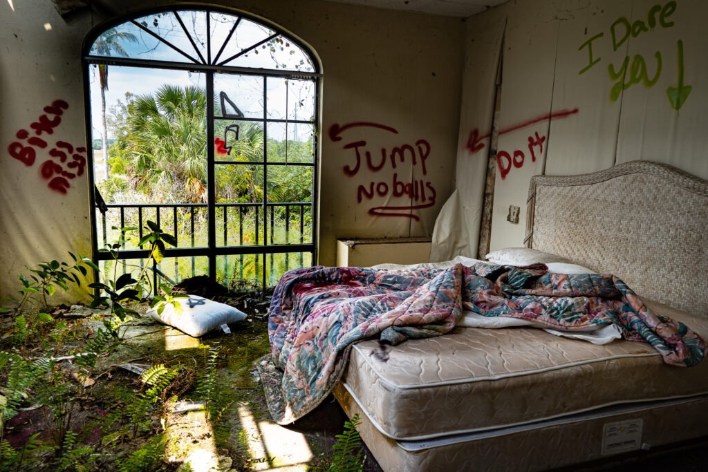 Inside a decayed hotel room at the abandoned Port of the Islands Resort in Naples, Florida. A disheveled bed with a floral comforter sits amid overgrown plants creeping through the broken window. Graffiti on the walls, including ominous messages like 'Jump' and 'I Dare You,' add to the eerie atmosphere. This former Everglades resort, now a hotspot for urban explorers, showcases nature reclaiming the forgotten space.