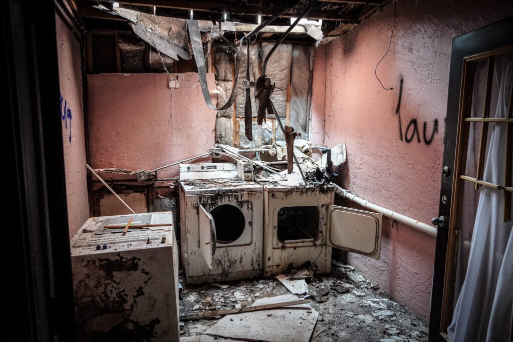 Abandoned laundry room at the demolished Port of the Islands Hotel in Naples, FL, featuring two rusted, open-door washing machines surrounded by debris and fallen ceiling panels. The pink walls, marked with graffiti, contrast with the exposed pipes and insulation hanging from the damaged ceiling. This image captures the scene of decay and neglect prior to the hotel's demolition.