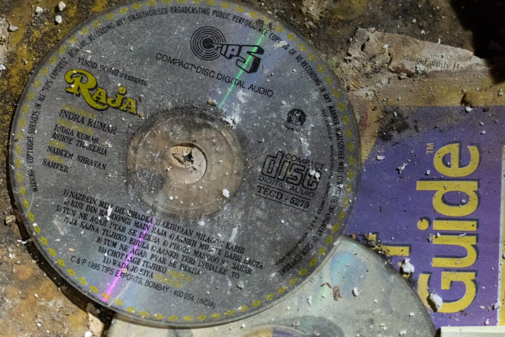 Close-up of a dusty and weathered compact disc (CD) labeled 'Raja,' featuring names like Indra Kumar, Nadeem-Shravan, and Sameer, found at the abandoned Port of the Islands Hotel in Naples, FL, prior to its demolition. The CD is partially covered in debris and lies atop a purple-and-yellow guidebook, capturing a piece of history and decay from the once-active hotel.