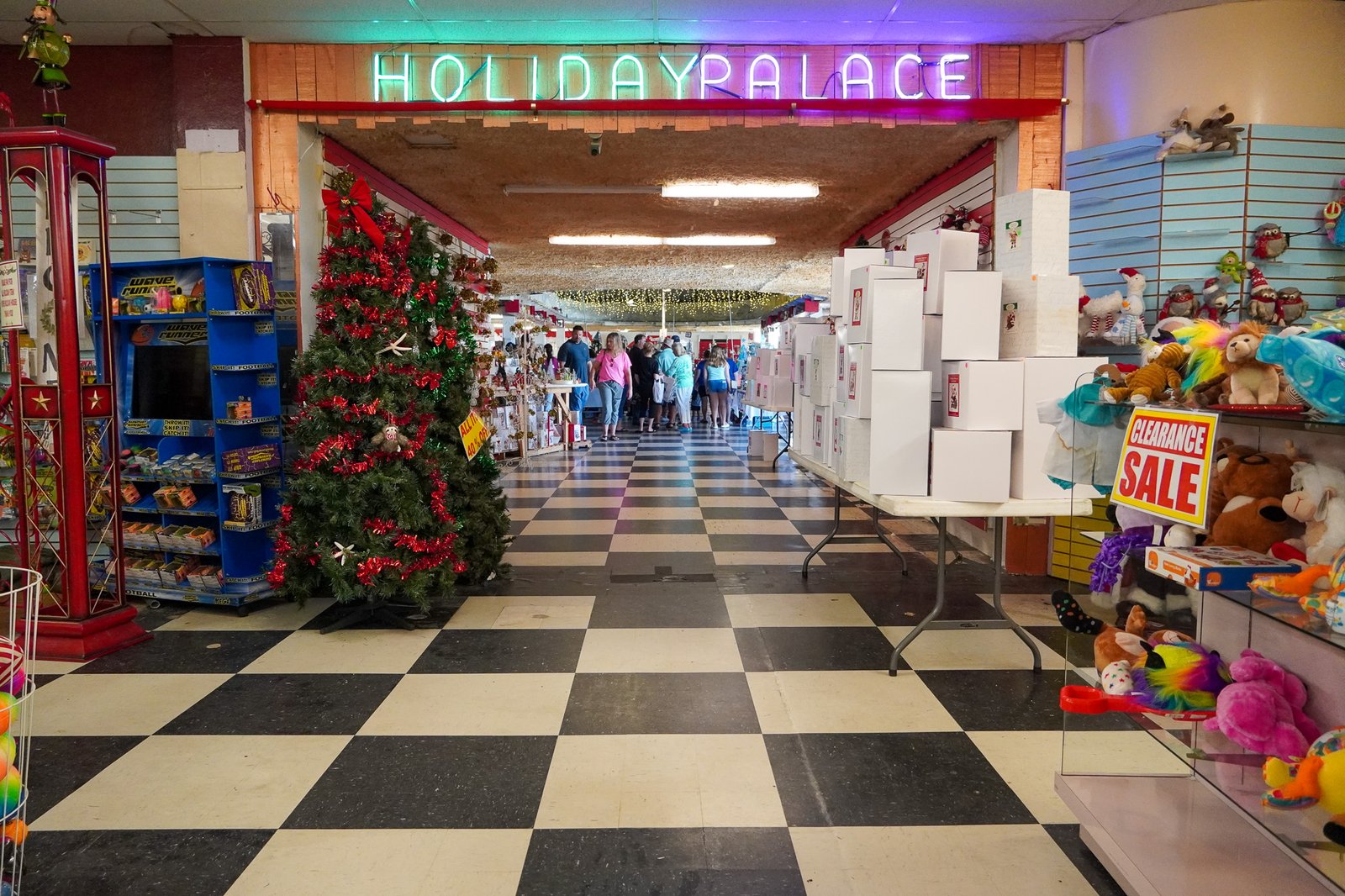 Interior view of the Shell Factory's Holiday Palace in Florida, highlighting a Christmas tree adorned with red tinsel and bows. The festive scene includes holiday decorations, clearance sale signs, and toy displays. Shoppers browse in the background under a neon sign reading 'HOLIDAY PALACE,' all set against a checkered black and white floor. This image captures the vibrant holiday shopping atmosphere at the iconic Shell Factory.