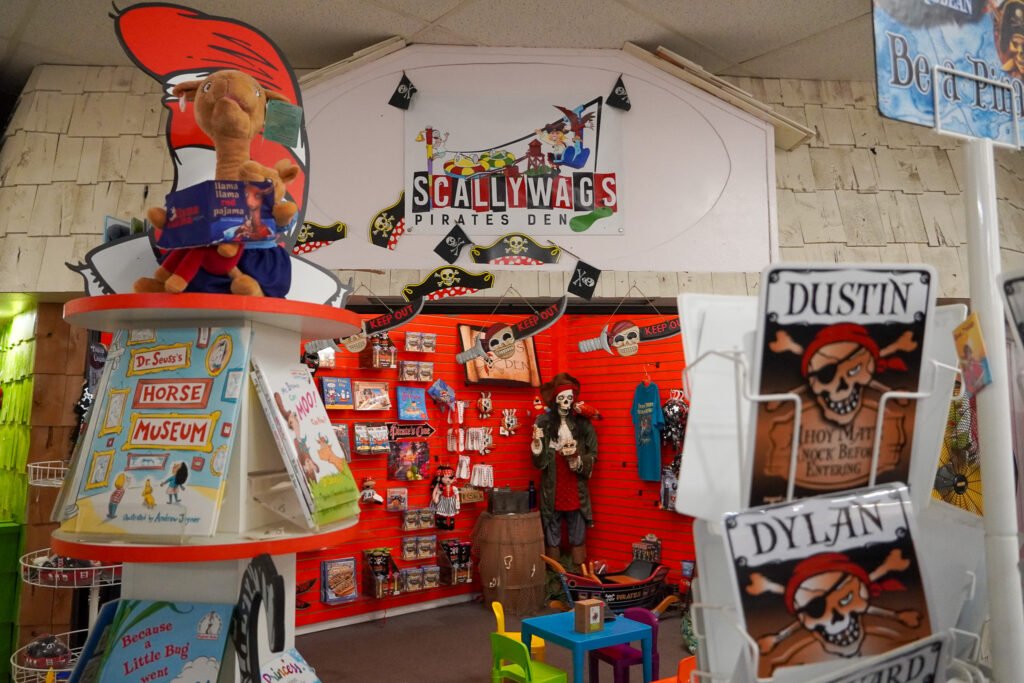 Scallywags Pirates Den inside the Shell Factory & Nature Park in Fort Myers, Florida. This pirate-themed children's area features colorful displays, a life-sized pirate mannequin, themed books, and novelty items. The store offers a variety of pirate-themed souvenirs, personalized name signs, and interactive decor for young adventurers.