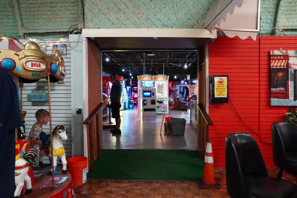 Entrance to the arcade at the Shell Factory & Nature Park in Fort Myers, Florida. The doorway leads into a dimly lit game room filled with flashing lights from various arcade machines. In the foreground, a child rides a small carousel while a man stands inside the arcade. The walls are decorated in a mix of red paneling and white slats, with a nostalgic, vintage feel. A traffic cone and caution sign are placed near the entrance, indicating maintenance or safety measures.