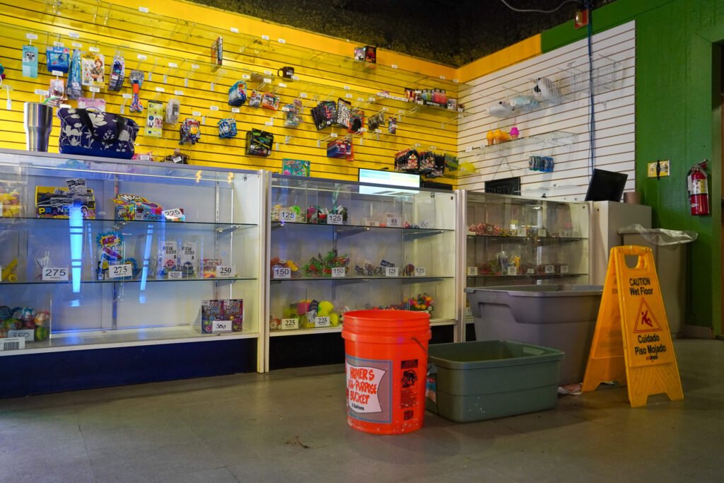 The prize redemption area inside the arcade at the Shell Factory & Nature Park in Fort Myers, Florida. Glass display cases contain small toys, candy, and other arcade prizes, while additional prizes hang on a yellow slat wall. Some shelves appear sparse. A bright orange bucket labeled 'Owner's Storage Bucket' sits on the floor, along with a green plastic tub, seemingly placed to catch water from a leak. A yellow 'Caution: Wet Floor' sign is also present, indicating possible maintenance issues.