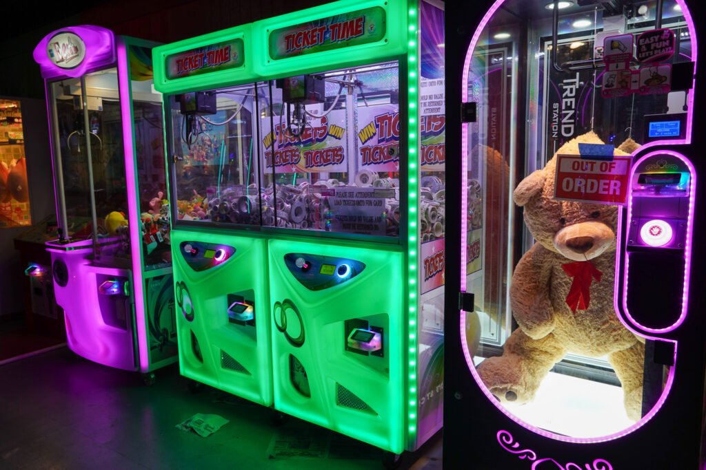 "A row of illuminated arcade claw machines at the Shell Factory & Nature Park in Fort Myers, Florida. The machines glow in vibrant shades of green, purple, and pink. One machine, labeled 'Ticket Time,' is filled with rolls of arcade tickets. Another machine features a giant plush teddy bear but is marked 'Out of Order' with a sign taped to its glass. The dimly lit arcade setting enhances the neon lighting, creating a nostalgic gaming atmosphere.