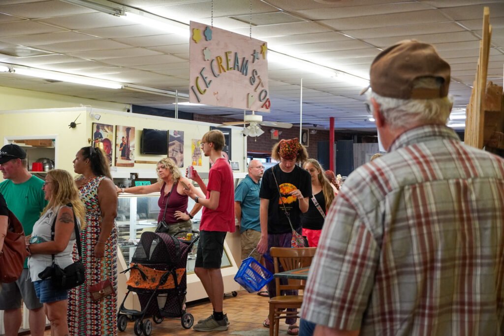 A line of people waiting at the Ice Cream Stop, an ice cream parlor inside the Shell Factory & Nature Park in Fort Myers, Florida. The crowd includes individuals of various ages, some holding shopping bags and others pushing strollers. A faded and slightly damaged 'Ice Cream Stop' sign hangs from the ceiling, adding to the worn atmosphere of the space. The ceiling tiles and lighting fixtures show signs of age, reflecting the overall condition of the building, which was further impacted by Hurricane Ian.