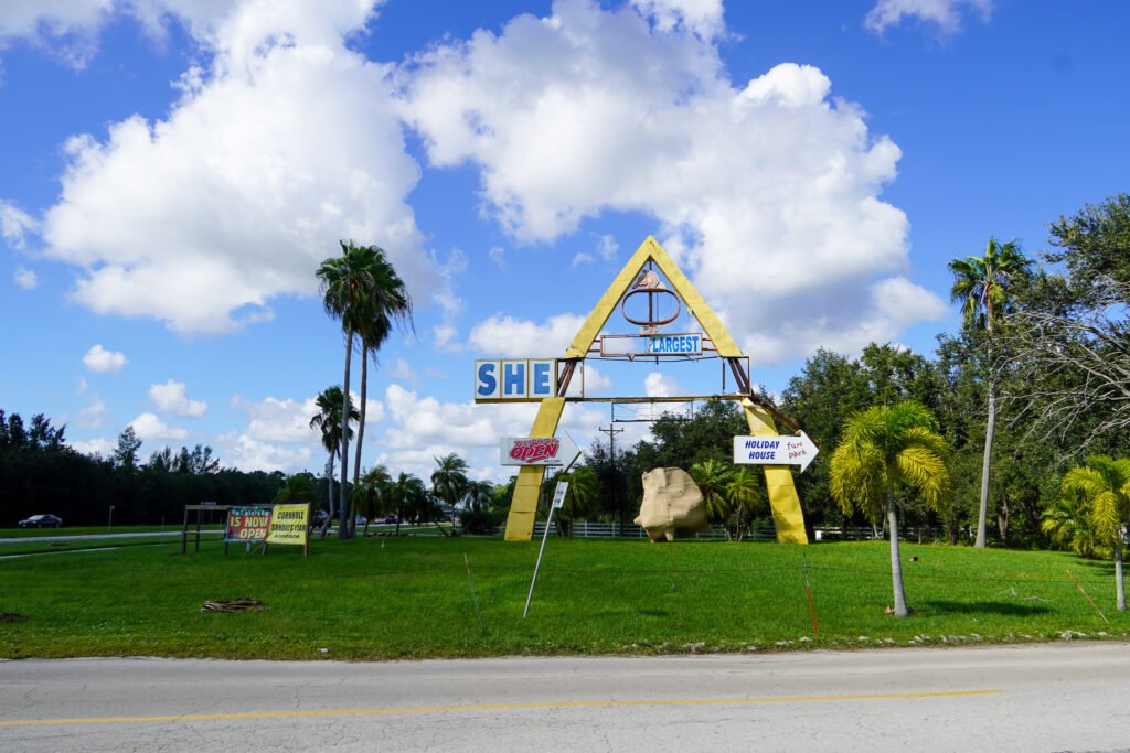 Iconic roadside entrance sign at the Shell Factory & Nature Park in Fort Myers, Florida, showing visible damage from Hurricane Ian. The large triangular structure with faded and broken lettering welcomes visitors to the attraction, known for its gift shop, wildlife park, and family-friendly activities. Lush palm trees and a bright blue sky provide a classic Florida backdrop.