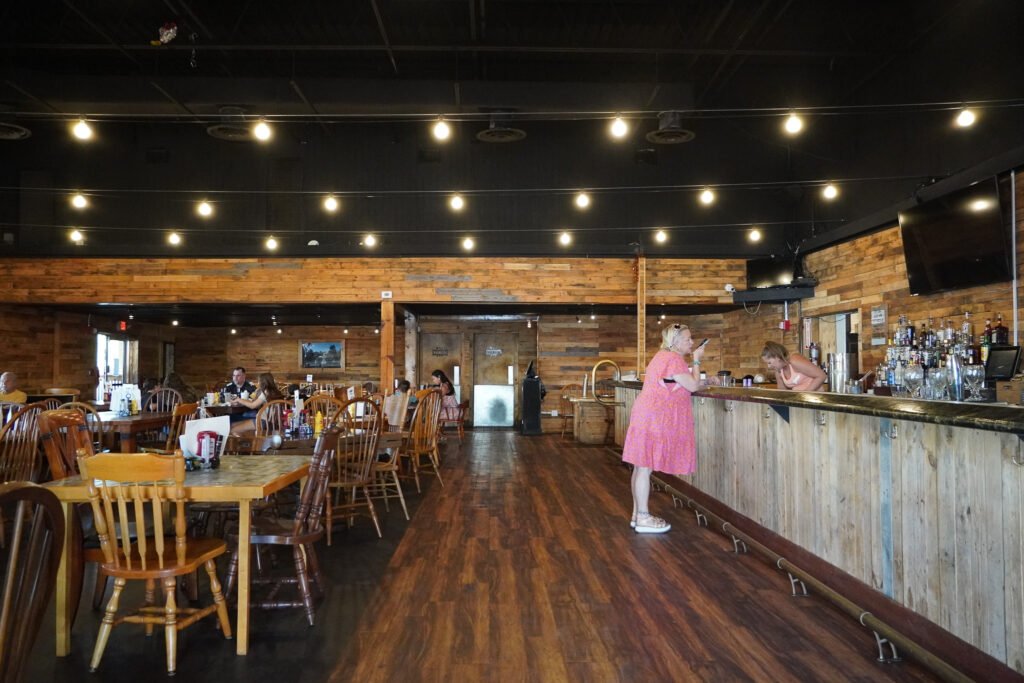 The interior of the restaurant at the Shell Factory & Nature Park in Fort Myers, Florida, on its final day before closing. With rustic wooden décor, string lights, and a nearly empty dining area, the restaurant is about to close for the last time. A woman in a pink dress speaks to the bartender at the long wooden bar, while a few remaining customers sit at tables. The Shell Factory, a longtime roadside attraction featuring a gift shop, arcade, and nature park, shut down in 2024 after decades in operation.