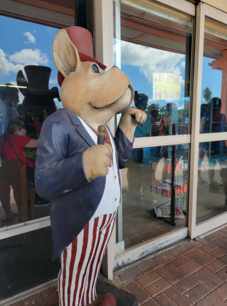 Whimsical anthropomorphic bulldog statue dressed as Uncle Sam outside the Shell Factory & Nature Park in Fort Myers, Florida. The patriotic dog figure wears a blue jacket, red-and-white striped pants, and a red top hat while holding a small cane. The Shell Factory, a popular roadside attraction, features unique souvenirs, nature exhibits, and nostalgic Americana decor.