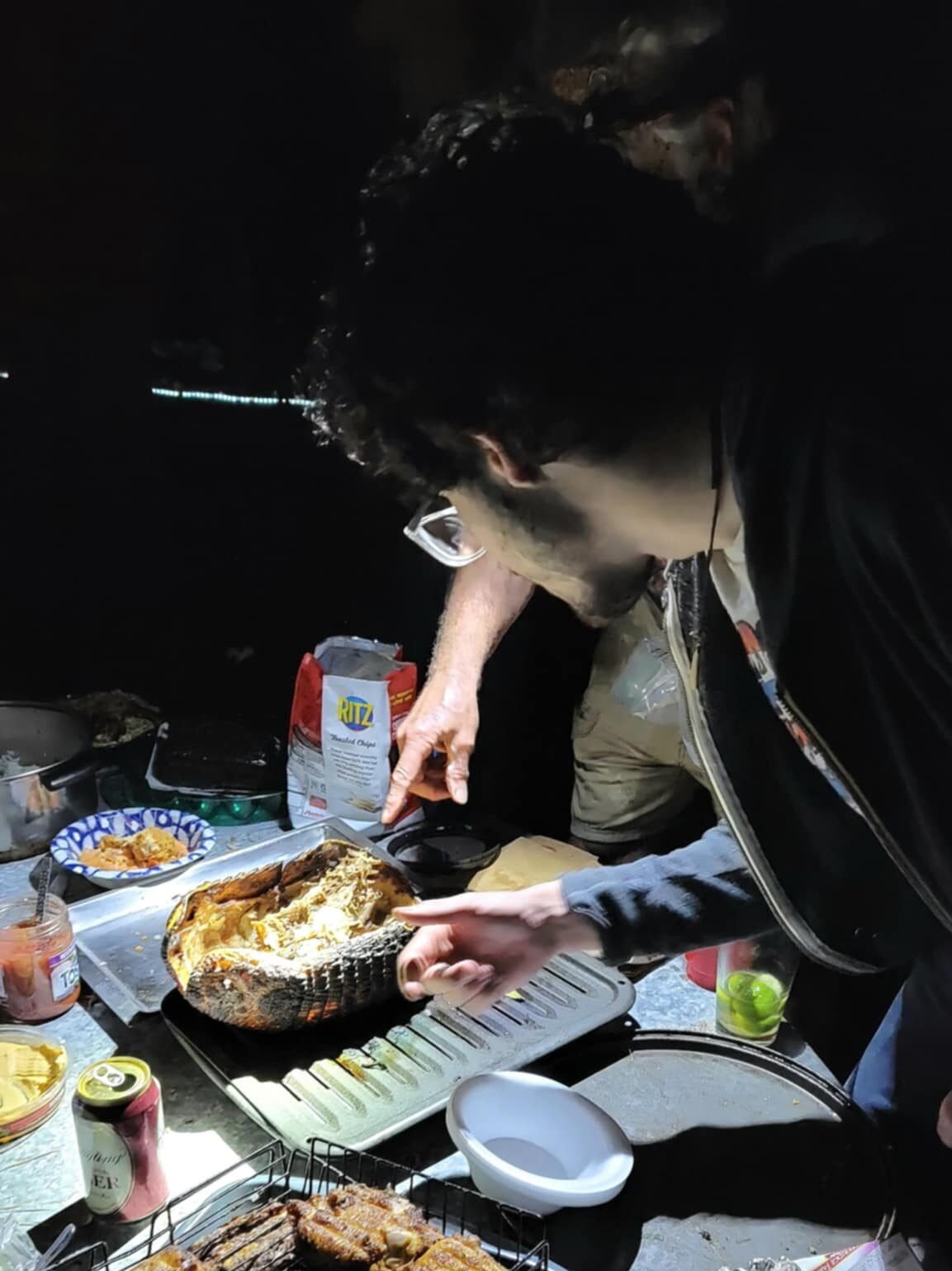 Outdoor gathering at Sleeze Weazel's 75th birthday party in Gainesville, Florida, featuring the infamous annual 'mystery meat' tradition. This year's dish is a cooked armadillo, split open and served on a grill under dim lighting. Guests are seen inspecting and preparing the unusual delicacy, surrounded by camping-style cookware, a can of beer, and snack items like Ritz crackers. The atmosphere is rustic and adventurous, reflecting the eccentric charm of Sleeze Weazel's legendary events.