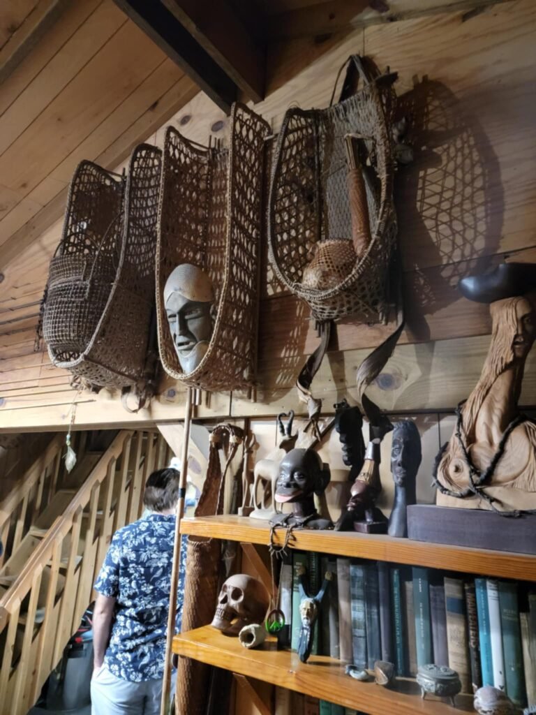 Inside the eclectic home of Sleeze Weazel in Gainesville, Florida, during his 75th birthday celebration. The rustic wooden interior features tribal masks, woven fish traps, carved wooden figurines, and antique books. A man in a Hawaiian shirt explores the space, adding to the mysterious and cultural ambiance of the collection. Unique artifacts and shadowed lighting create a museum-like atmosphere.