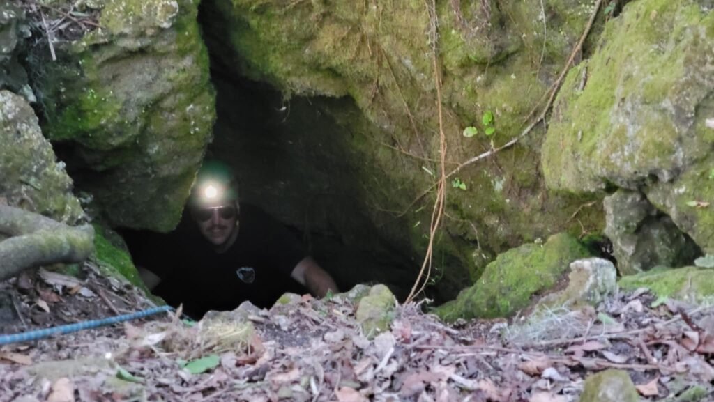 A Florida caver wearing a headlamp and sunglasses emerges from the entrance of a moss-covered cave. The rocky opening is surrounded by green foliage and vines, creating a natural and adventurous setting