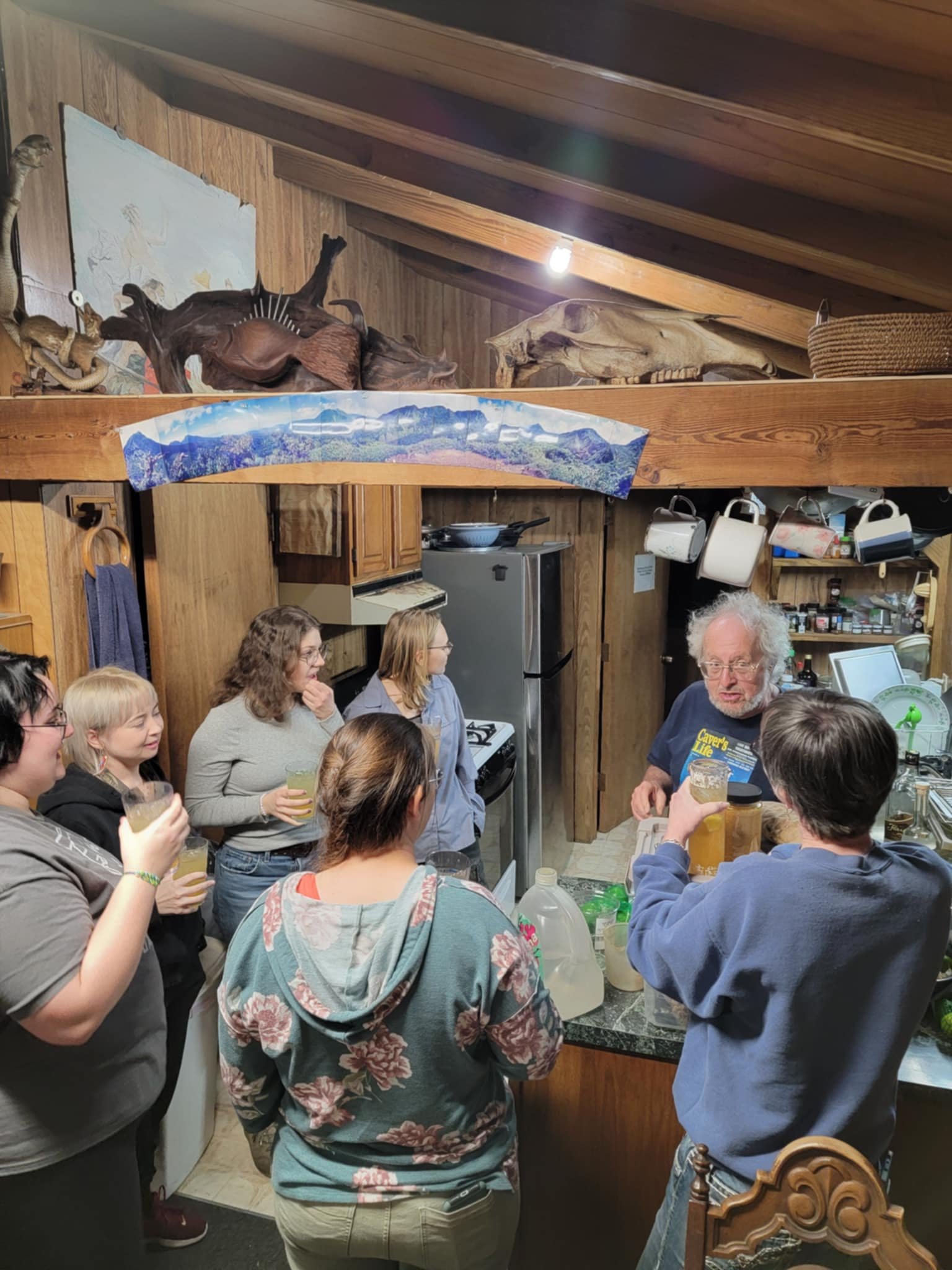 A group of people gathered in a rustic kitchen inside Sleeze Weazel's house during his birthday celebration. Sleeze Weazel, an infamous Gainesville figure and retired international landscape artist, is seen with white hair and a beard, wearing a 'Caver's Life' t-shirt while preparing drinks. The warm, wood-paneled interior features eclectic decorations, including animal skulls and carved artwork. Attendees, mostly young adults, are engaged in conversation and enjoying homemade beverages. Sleeze Weazel is also known as the designer of the butterfly garden at the Gainesville Natural History Museum.