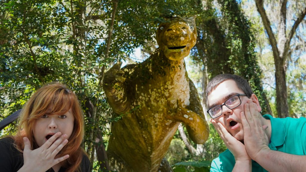 Two surprised visitors pose with an old dinosaur statue at Dunlawton Sugar Mill Gardens in Port Orange, Florida. This moss-covered statue is a relic from Bongoland, a defunct 1940s roadside attraction. Surrounded by lush greenery, the historic site blends Florida history with quirky remnants of vintage tourism, making it a hidden gem for visitors.