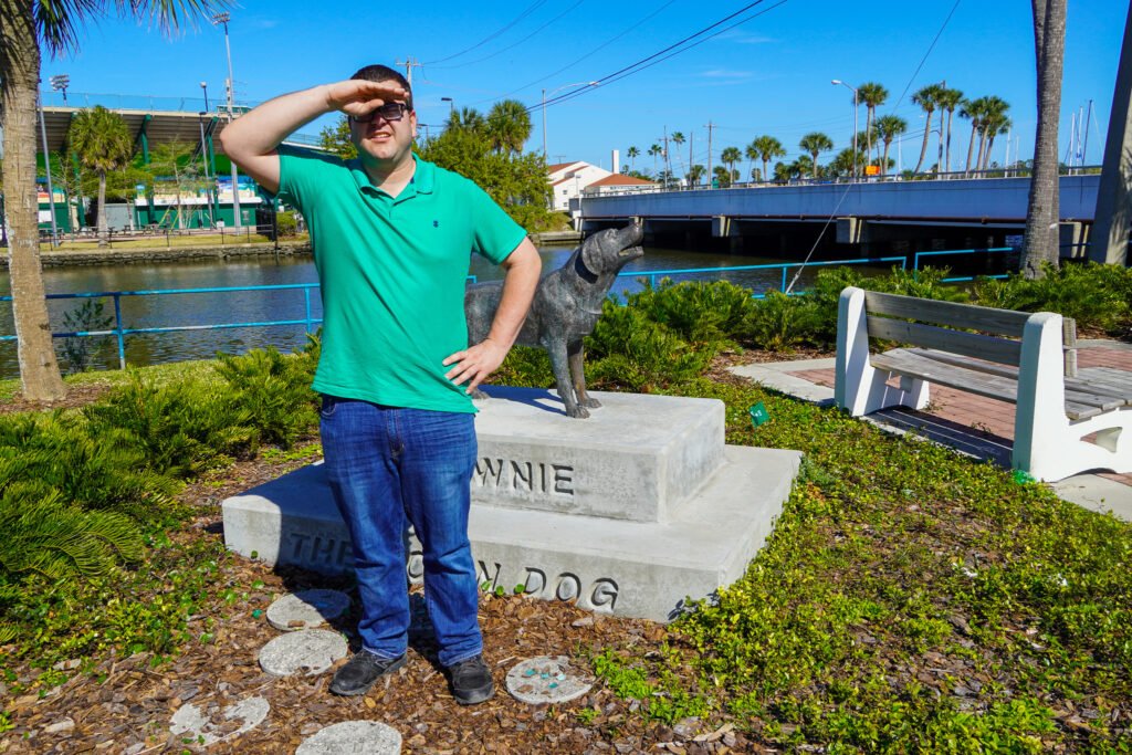 A visitor poses next to the Brownie the Town Dog Memorial Statue in Daytona Beach, Florida. This statue honors Brownie, the beloved stray dog who became the unofficial mascot of the city in the 1940s. Located near the Halifax River, the memorial celebrates Brownie's legacy as Daytona’s most famous canine resident, attracting tourists and dog lovers alike.