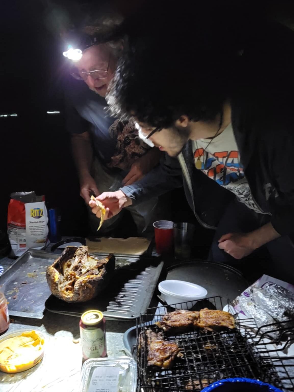 "Sleeze Weazel's 75th birthday party in Gainesville, Florida, featuring the infamous annual 'mystery meat' tradition. This year's dish is a roasted armadillo, served on a grill in an outdoor nighttime setting. A man wearing glasses and a graphic T-shirt is seen pulling meat from the armadillo with a Ritz cracker, while an older man with a headlamp watches. The rustic spread includes grilled meats, cheese dip, and cans of beer, adding to the eccentric and adventurous atmosphere of this legendary Gainesville gathering.